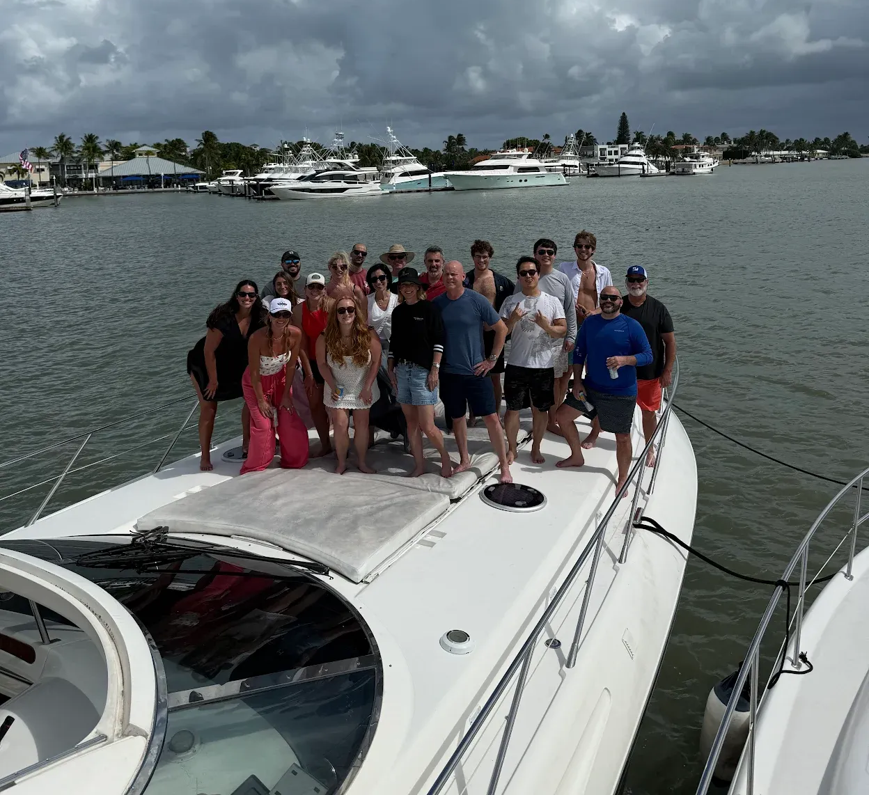 Group of people posing on a boat in a harbor. Cloudy sky, other boats docked in the background.