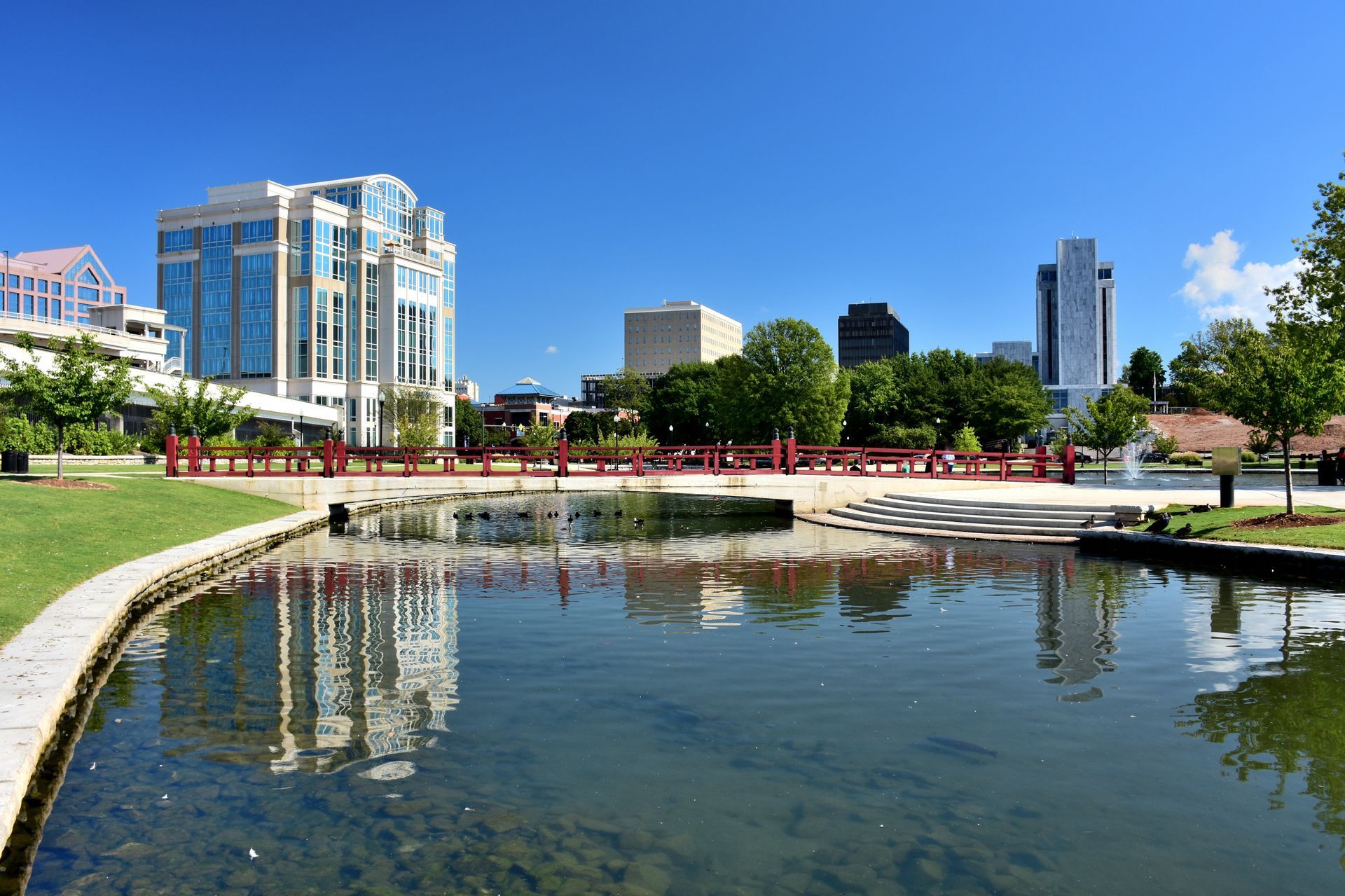 City park with water feature; buildings and blue sky reflected in the water.