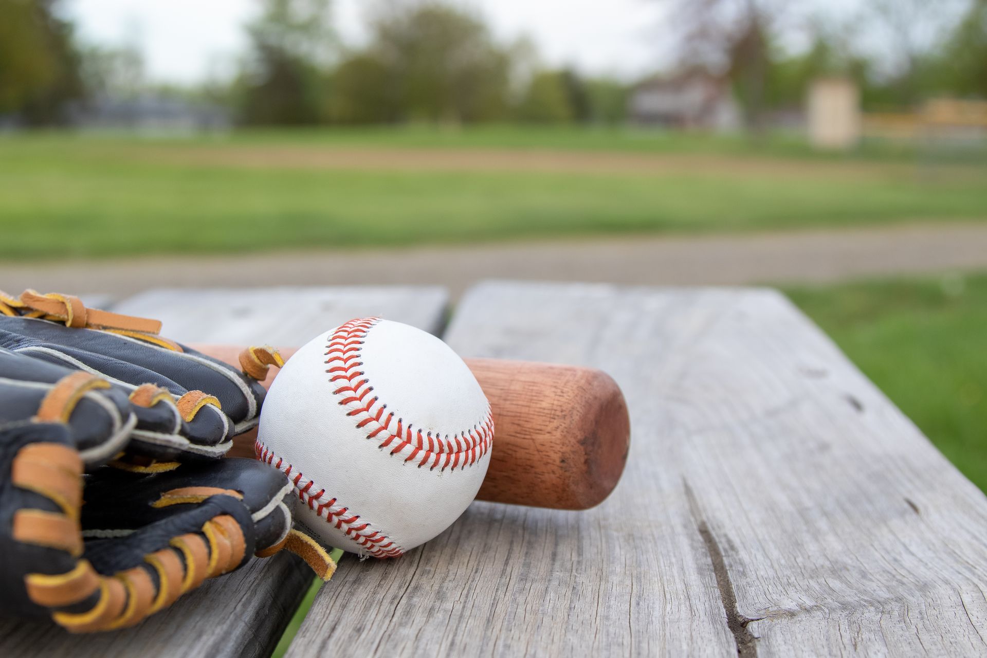 Baseball glove, ball, and bat on a weathered picnic table with a baseball field in the background.