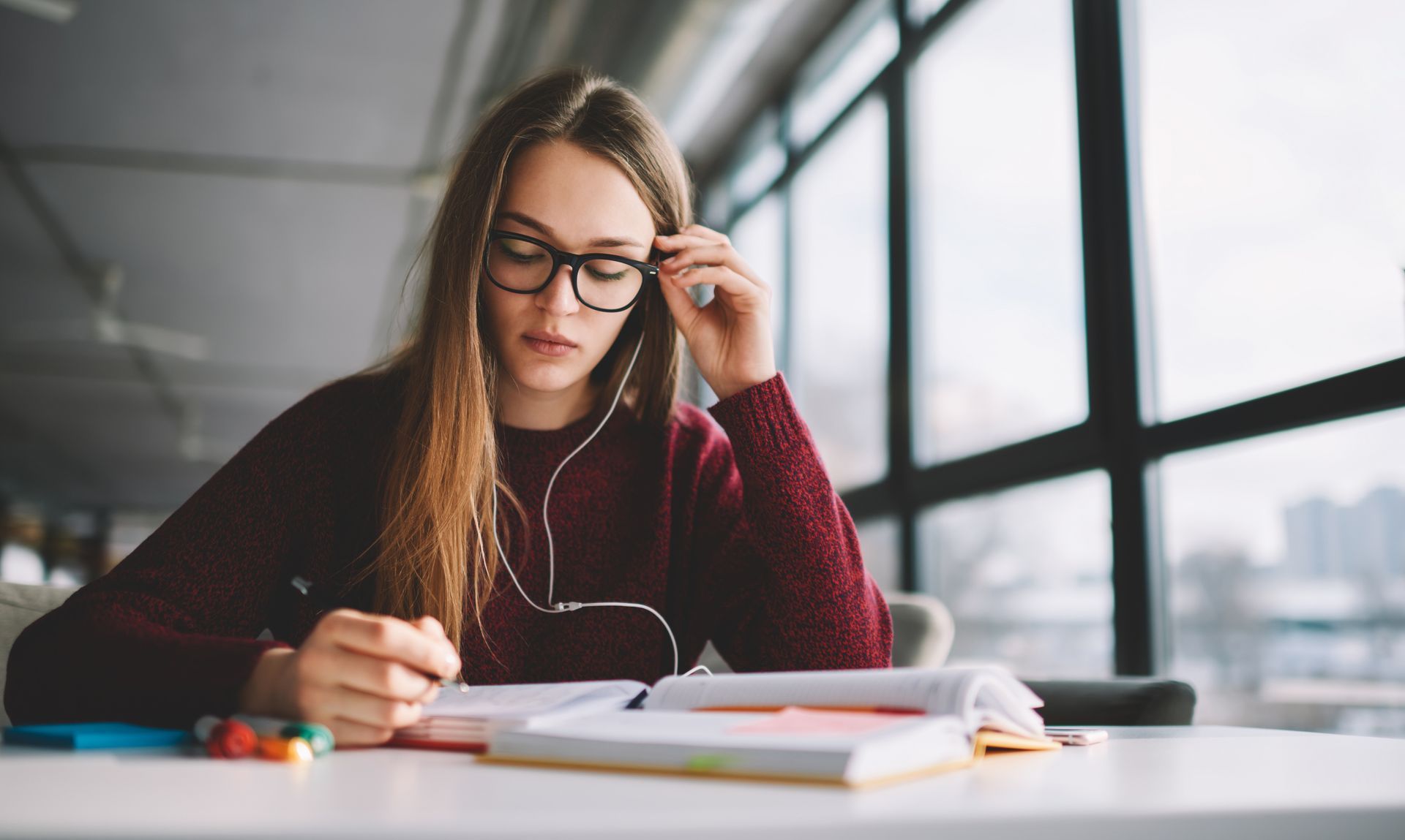 A young woman is sitting at a table wearing headphones and writing in a notebook.