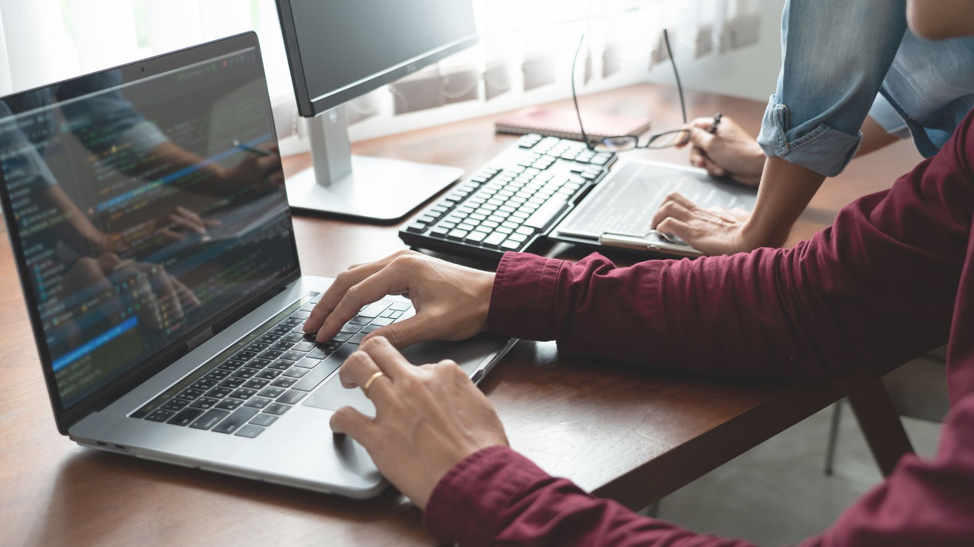 Person typing on laptop, others work nearby. Programming code displayed on screen, indoors.