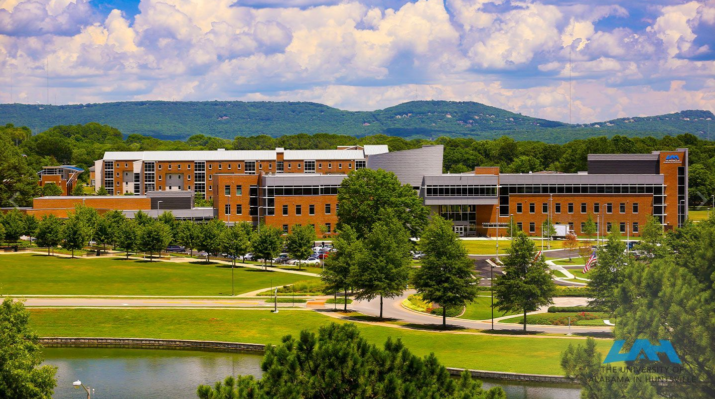 Large brick building with a modern design, on a green lawn by a lake, hills in the background. Sunny day.