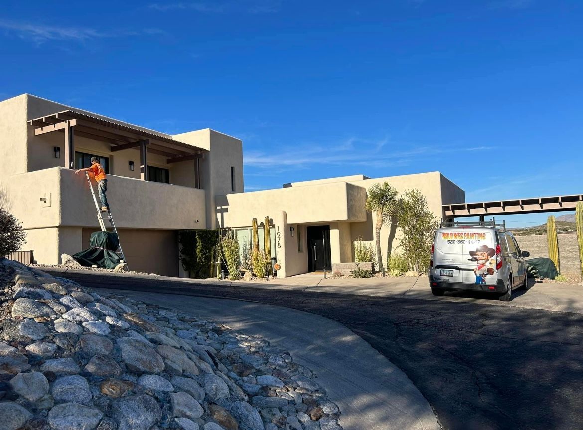 A technician on a ladder works on the roof of a beige house. A service van is parked nearby on a sunny day.