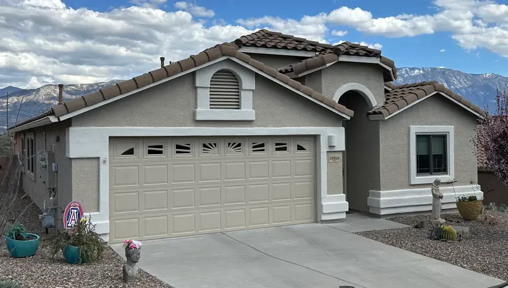 Tan stucco house with a brown tile roof, driveway, and mountain backdrop on a cloudy day.