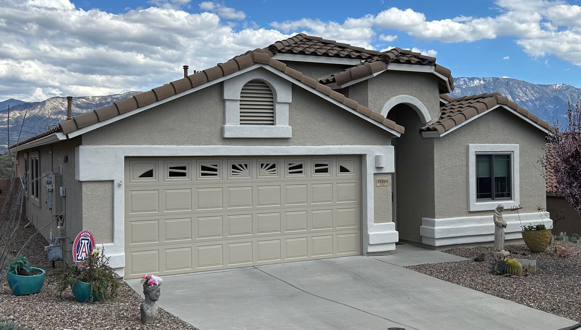 Tan stucco home with a beige garage door, arched entryway, and tile roof against a backdrop of mountains and a cloudy sky.
