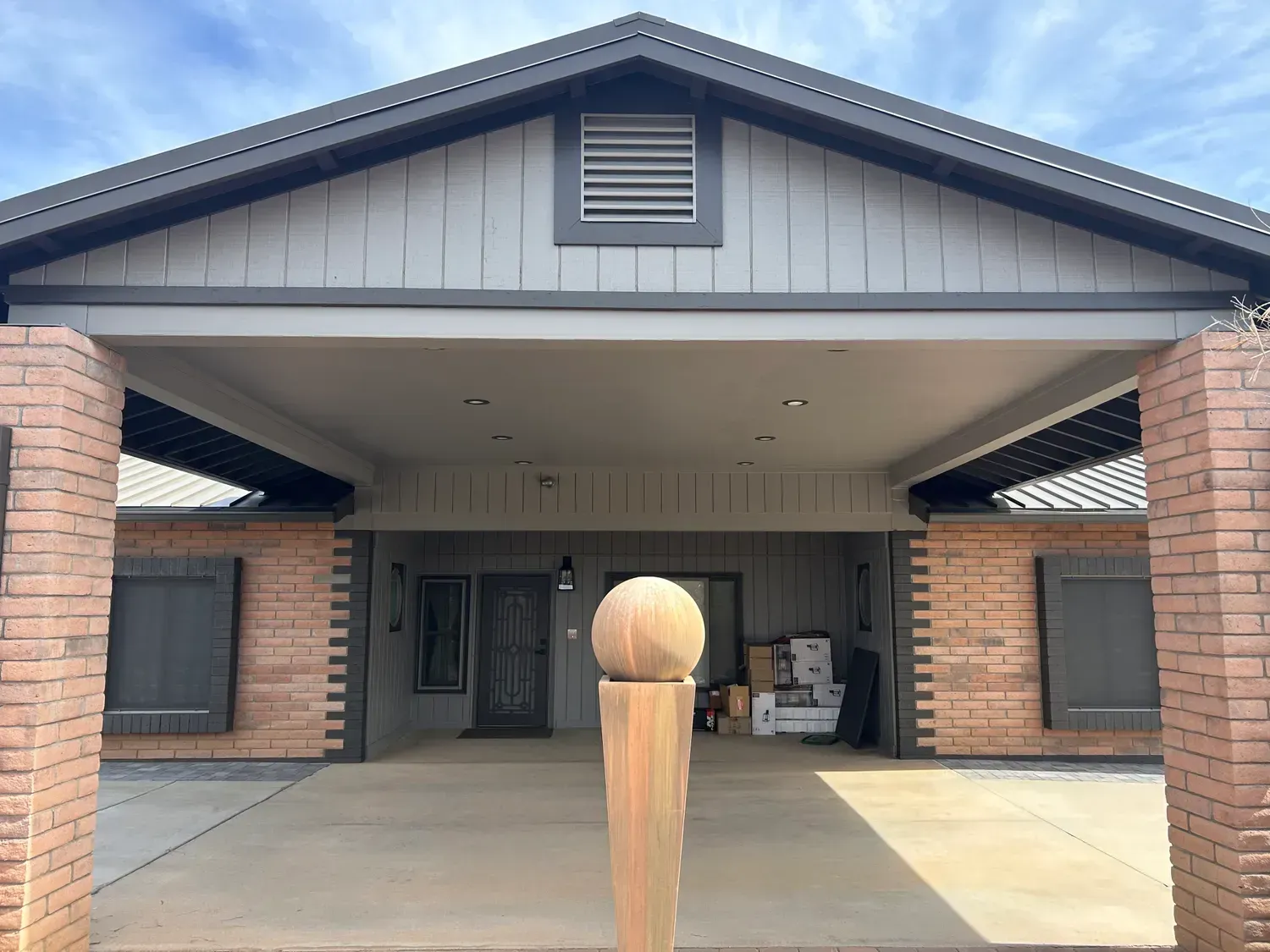 Exterior view of a building with a covered entrance, supported by brick pillars. The structure features gray siding
