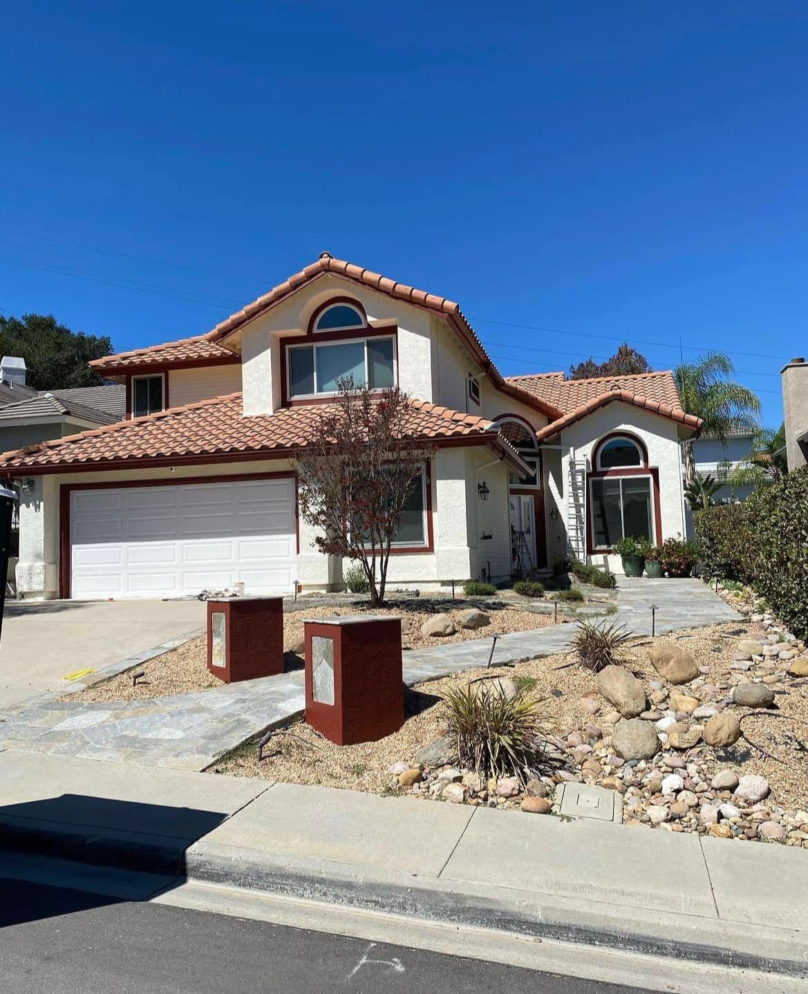 Two-story stucco house with red tile roof and garage. A winding walkway and front yard feature rocks and dry landscaping.