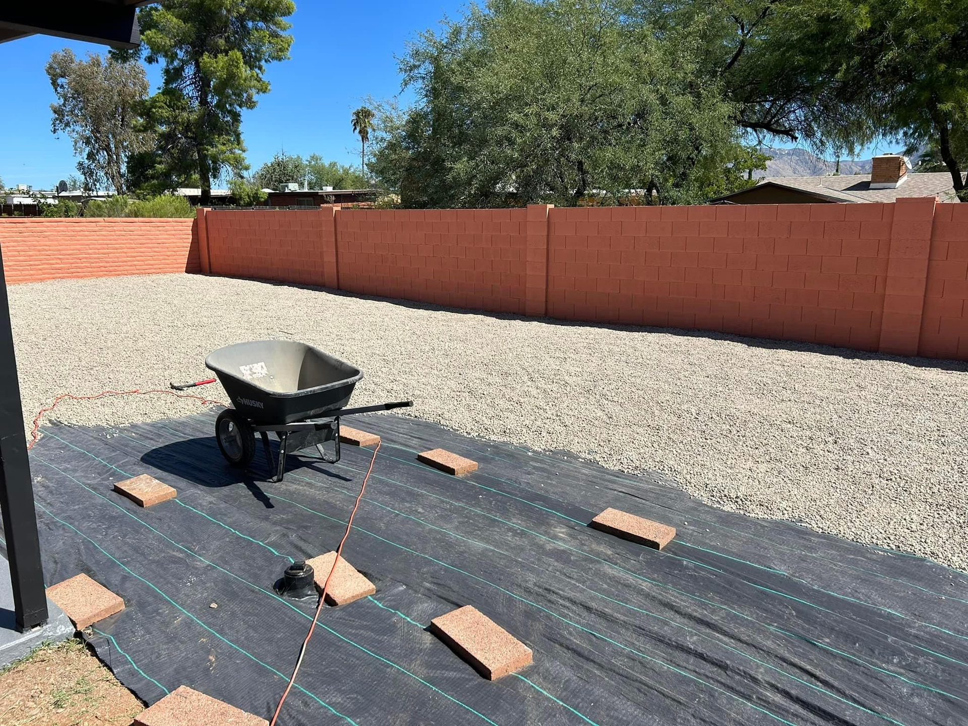 Backyard with a wheelbarrow on weed barrier, surrounded by gravel and a red wall. Bricks mark a planned garden area.