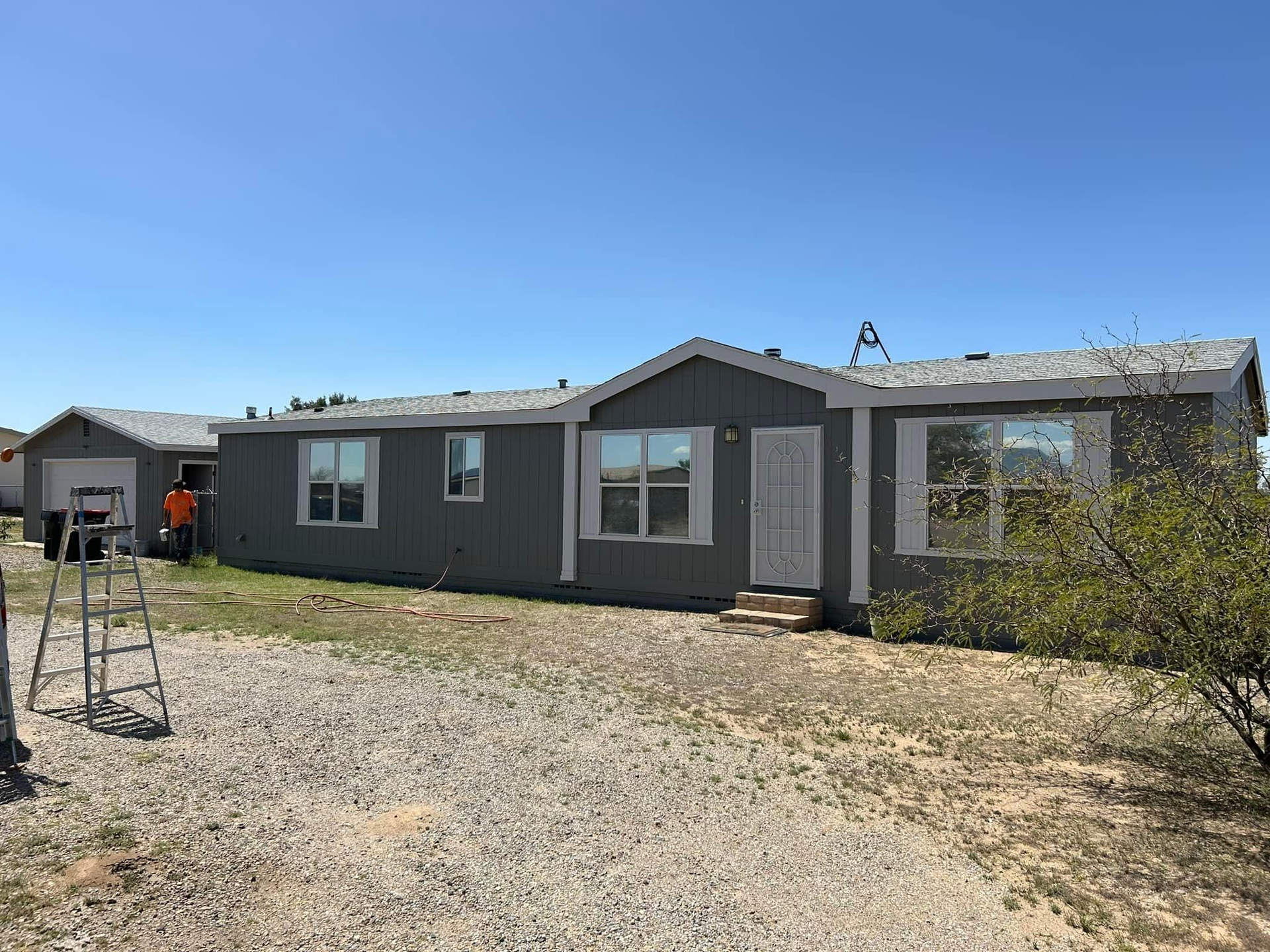 A gray, single-story house with a matching detached garage under a blue sky. A person is visible near the garage.