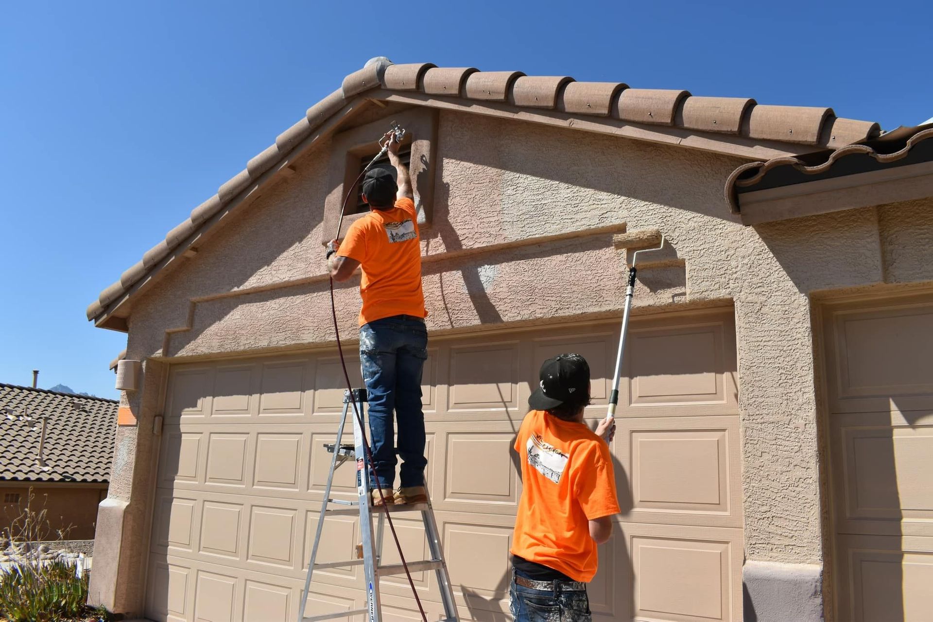 Two painters in orange shirts repaint a beige garage on a sunny day. One is on a ladder; the other uses a roller.