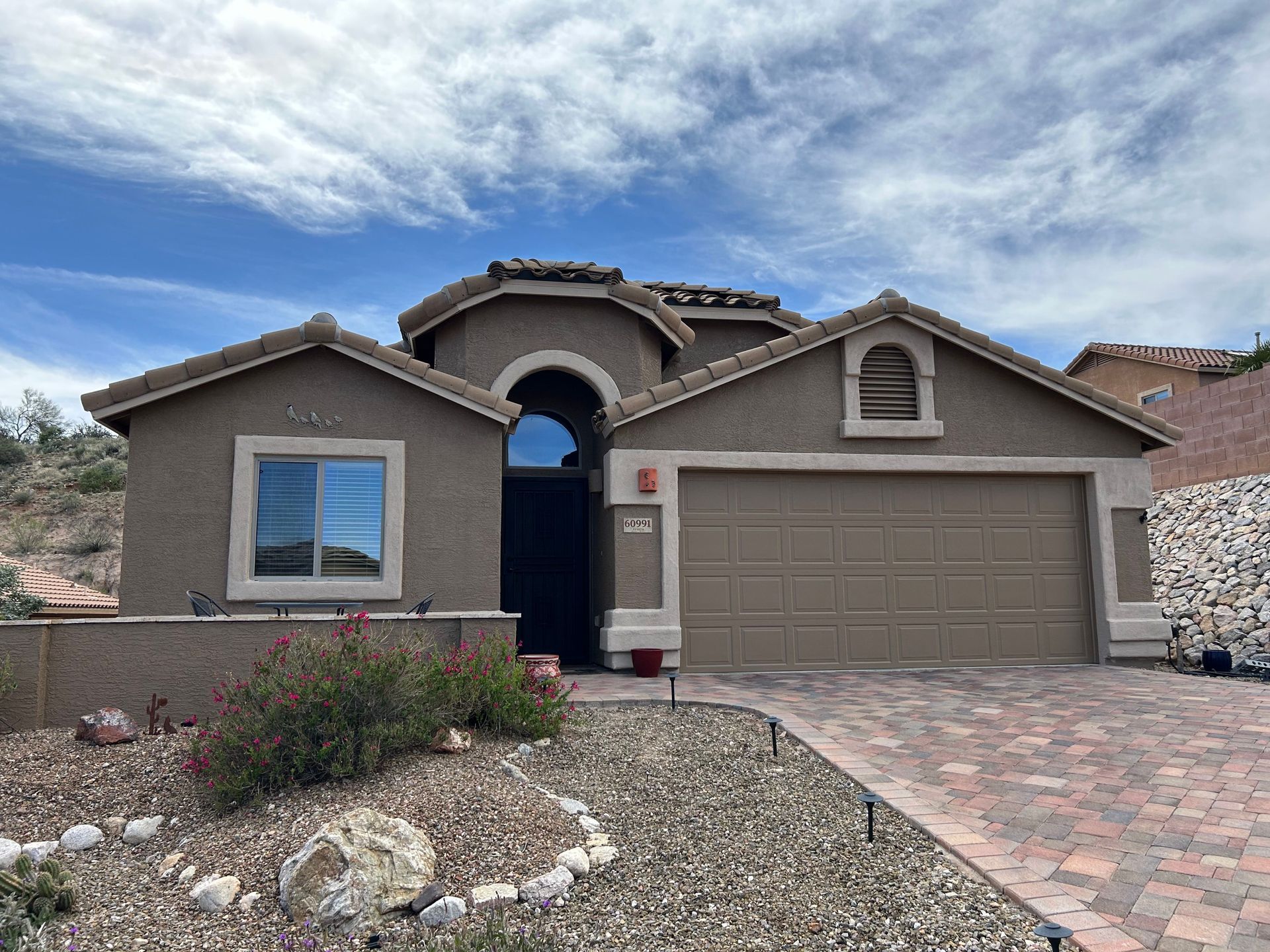 Tan stucco house with a brown garage door and brick driveway under a cloudy blue sky.  