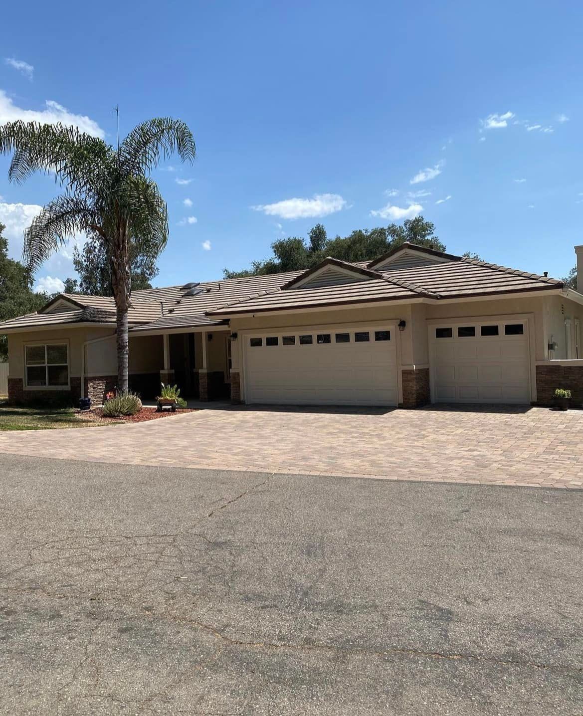 A single-story beige house with a three-car garage, brick driveway, and a palm tree against a blue sky.