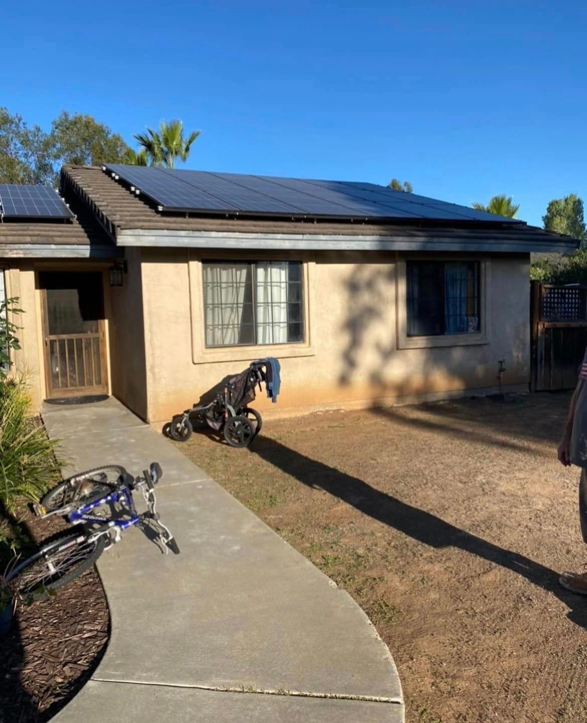 A one-story house with solar panels, a concrete walkway, and a bicycle in the foreground. Sunny day.