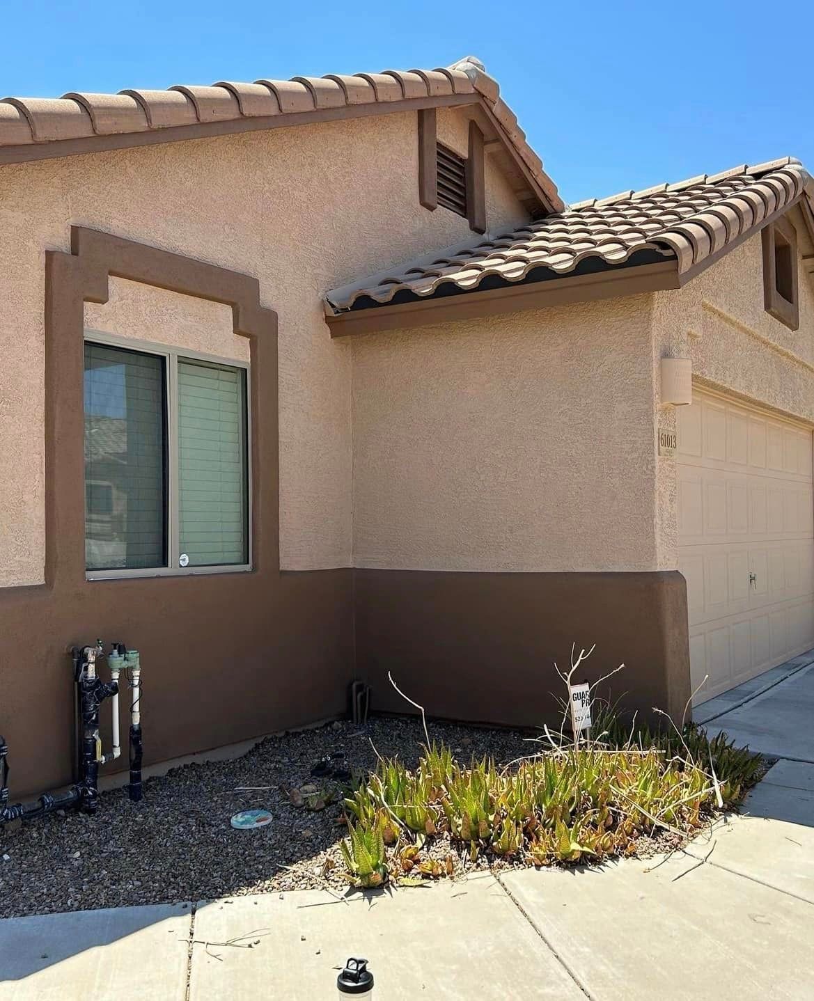 Exterior of a tan stucco house with a brown accent wall and a garage door, sunny day.