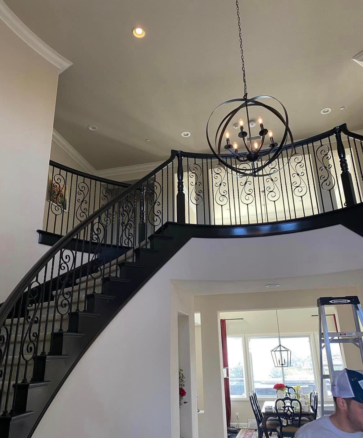 A grand foyer with a curved staircase and black wrought-iron railing. A large chandelier hangs from the high ceiling.