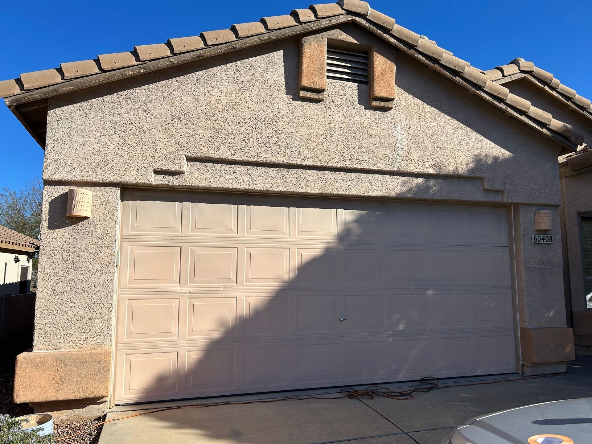 A beige garage door on a stucco house with brown roof tiles and a vent above. The garage door has a shadow on it.