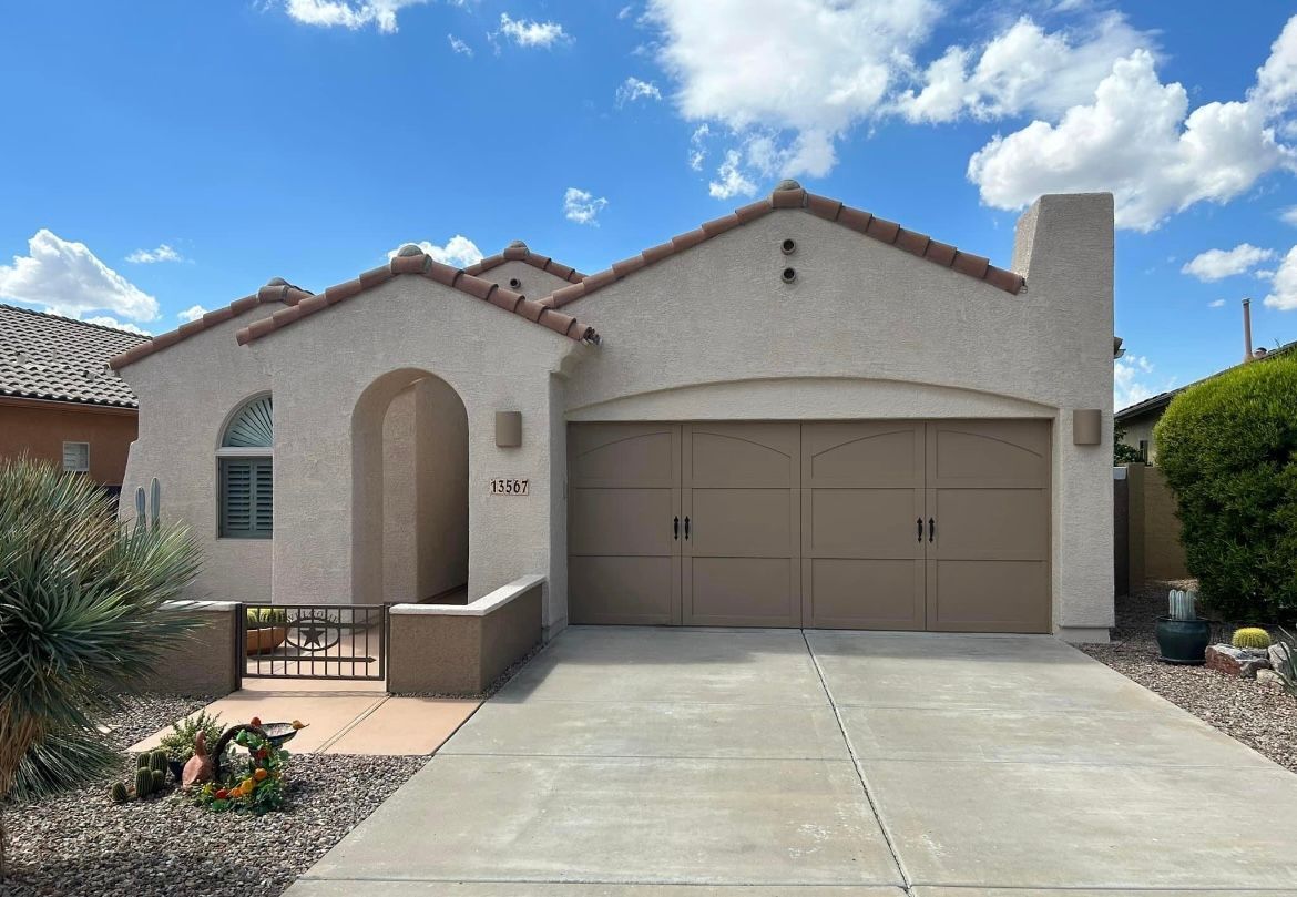 Tan stucco house with a brown garage door and arched entryway under a blue sky.