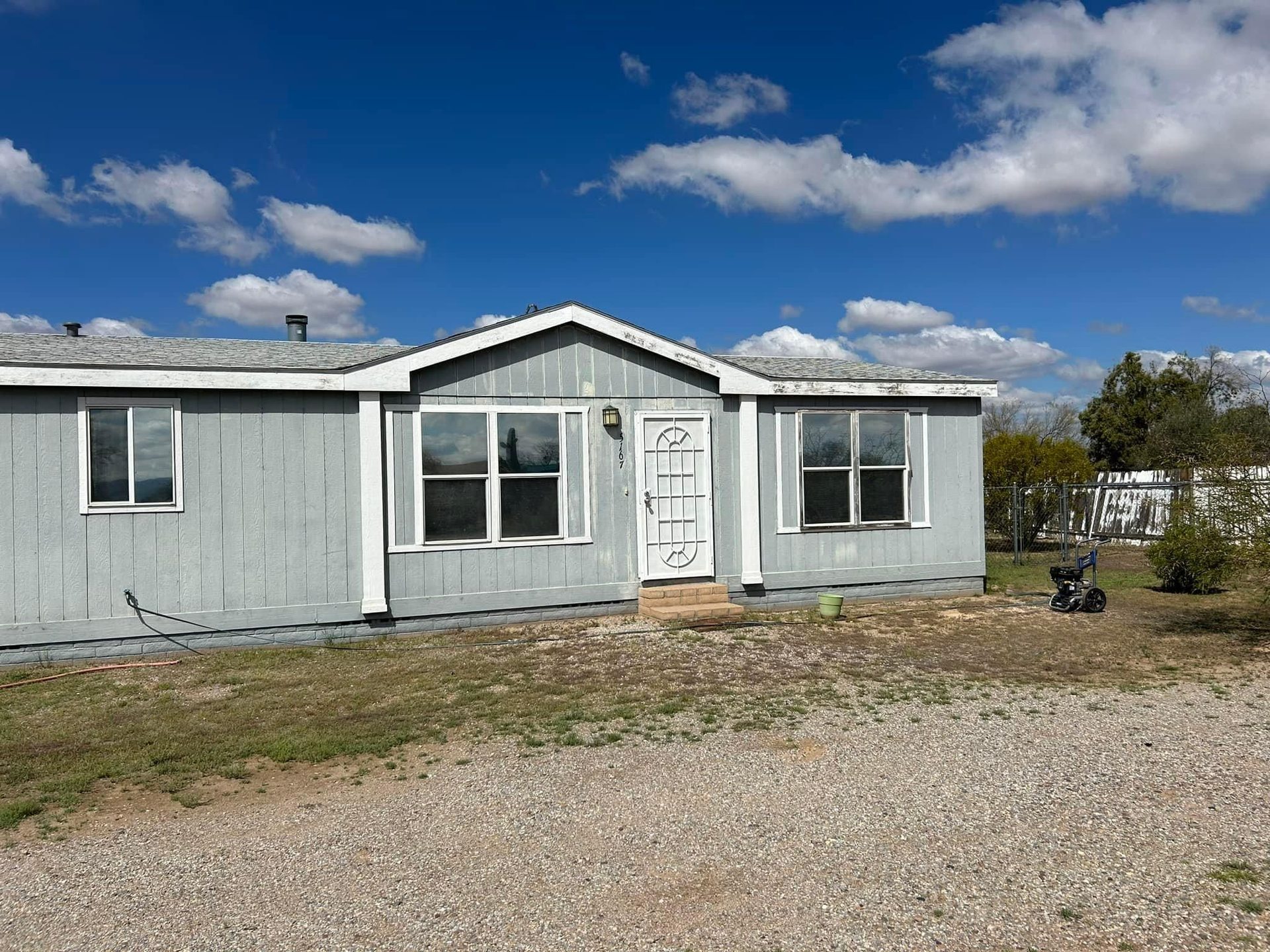 A light blue mobile home with white trim and a white front door sits on a gravel lot under a partly cloudy sky.