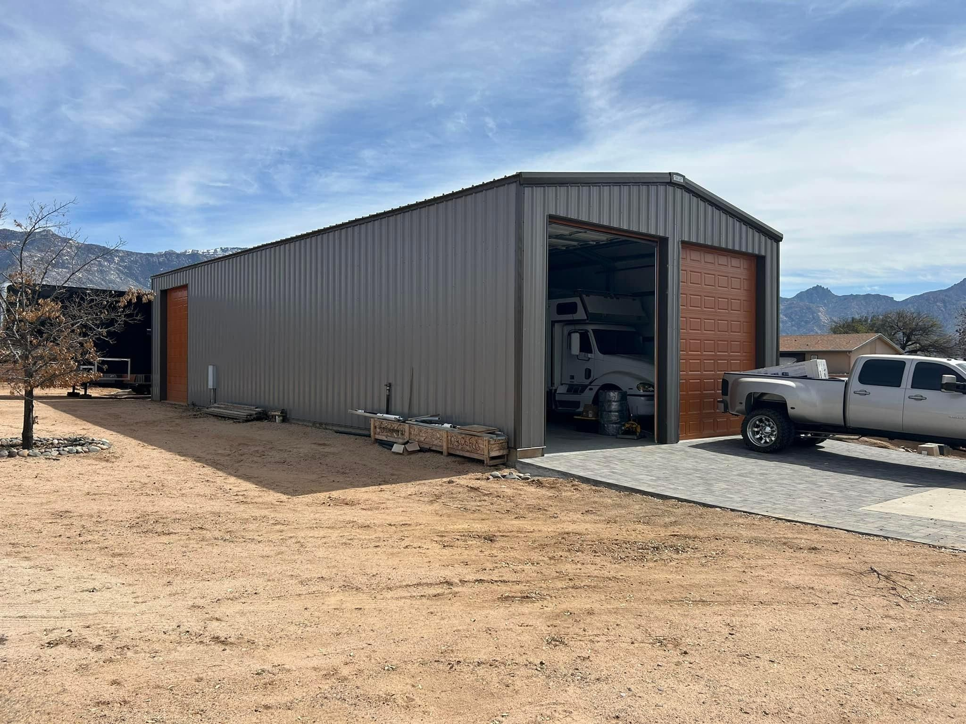 A gray metal shed with open bays, housing a camper and truck, sits on a gravel driveway under a partly cloudy sky.