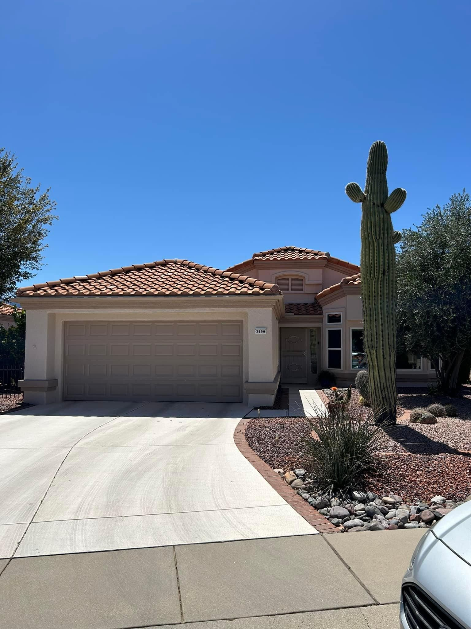 A beige house with a curved driveway, tile roof, and large cactus in front, under a clear blue sky.