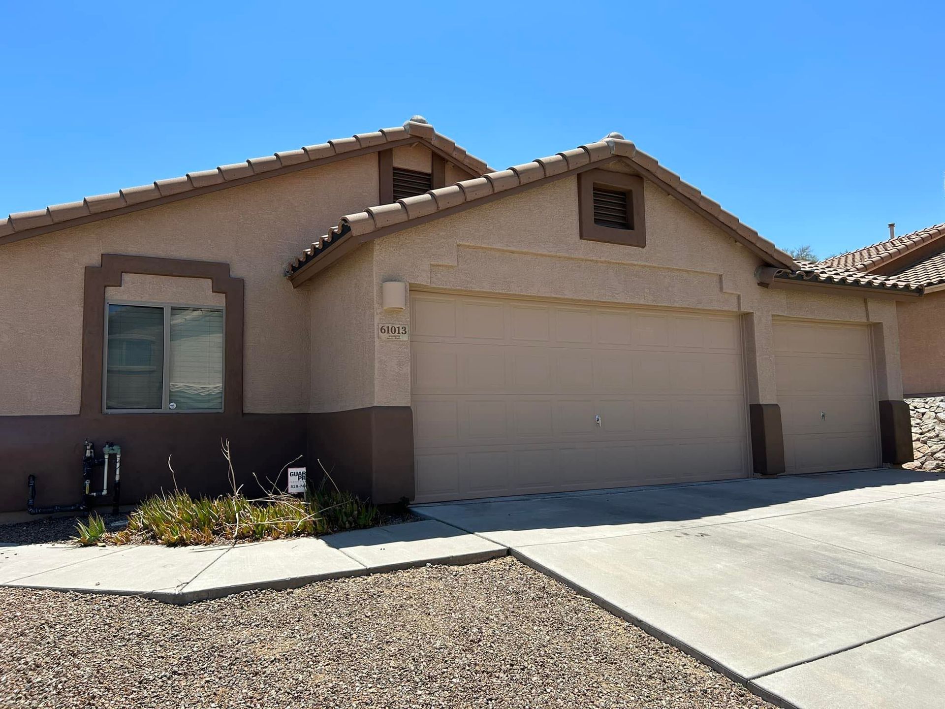 Tan stucco house with three-car garage, brown trim, and clear blue sky. A small front yard with a few plants is visible.