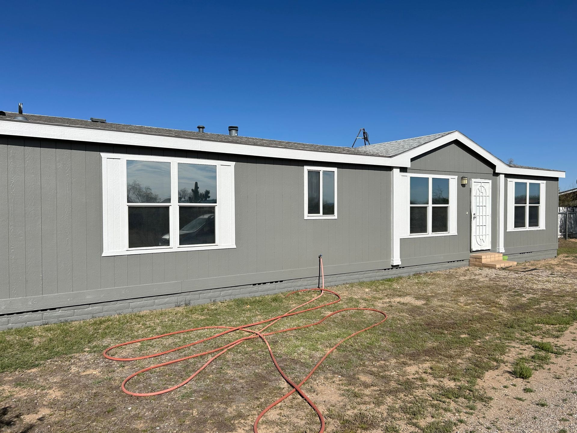 Gray mobile home with white trim and a blue sky. An orange hose lies on the patchy grass in front.