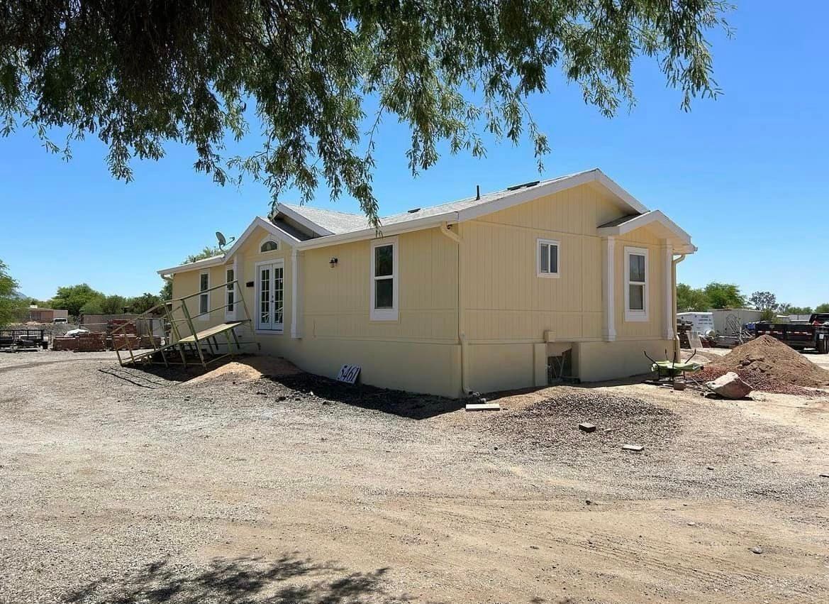 A yellow house with a gray roof sits on a gravel lot under a clear blue sky. Trees partially frame the view.