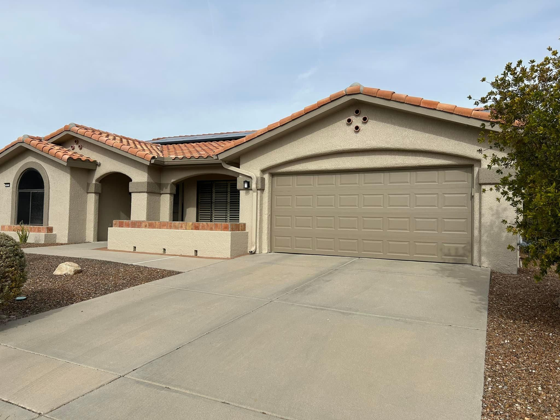 Tan stucco ranch-style home with a brown garage door, red-tiled roof, and a concrete driveway on a sunny day.