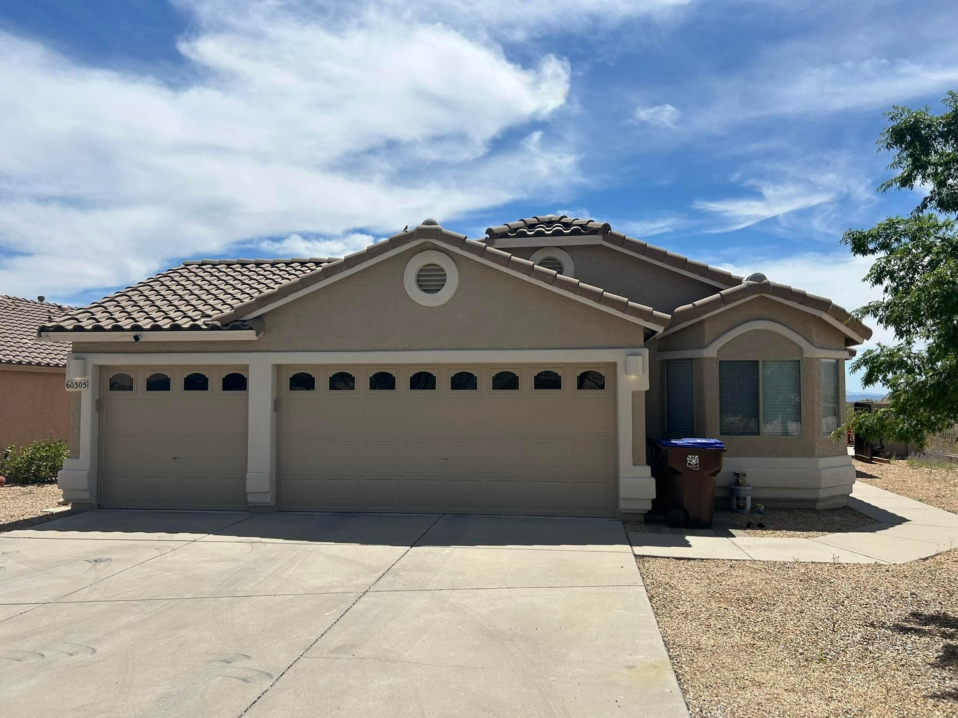 Tan-colored suburban house with a two-car garage, driveway, and gravel landscaping under a blue sky.