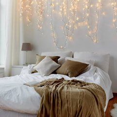 Bedroom with fairy lights on the wall behind the bed, layered cushions with throw rug draped over the end of the bed.