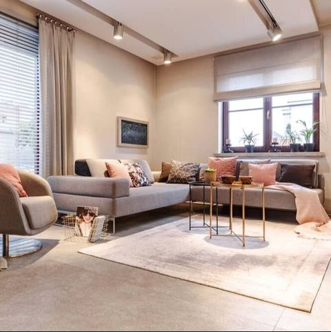 Neutral tone loungeroom, with open curtains showing venetians on one window, roman blind half way up on other window. Rug in front of couch with cushions on concrete floor.