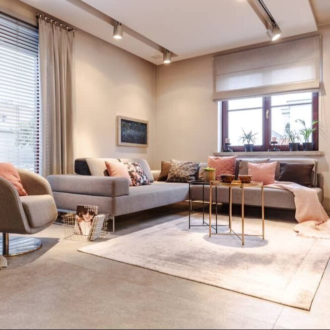 Neutral tone loungeroom, with open curtains showing venetians on one window, roman blind half way up on other window. Rug in front of couch with cushions on concrete floor.