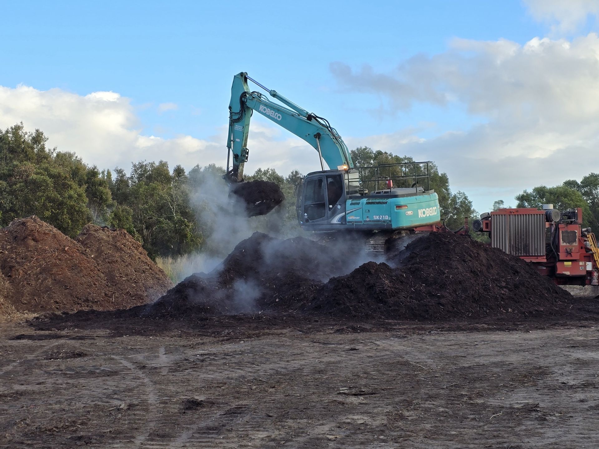 An excavator, mint green, moves a pile of dark compost outdoors, creating steam — Coastal Sand Soil & Mulch in Craignish, QLD
