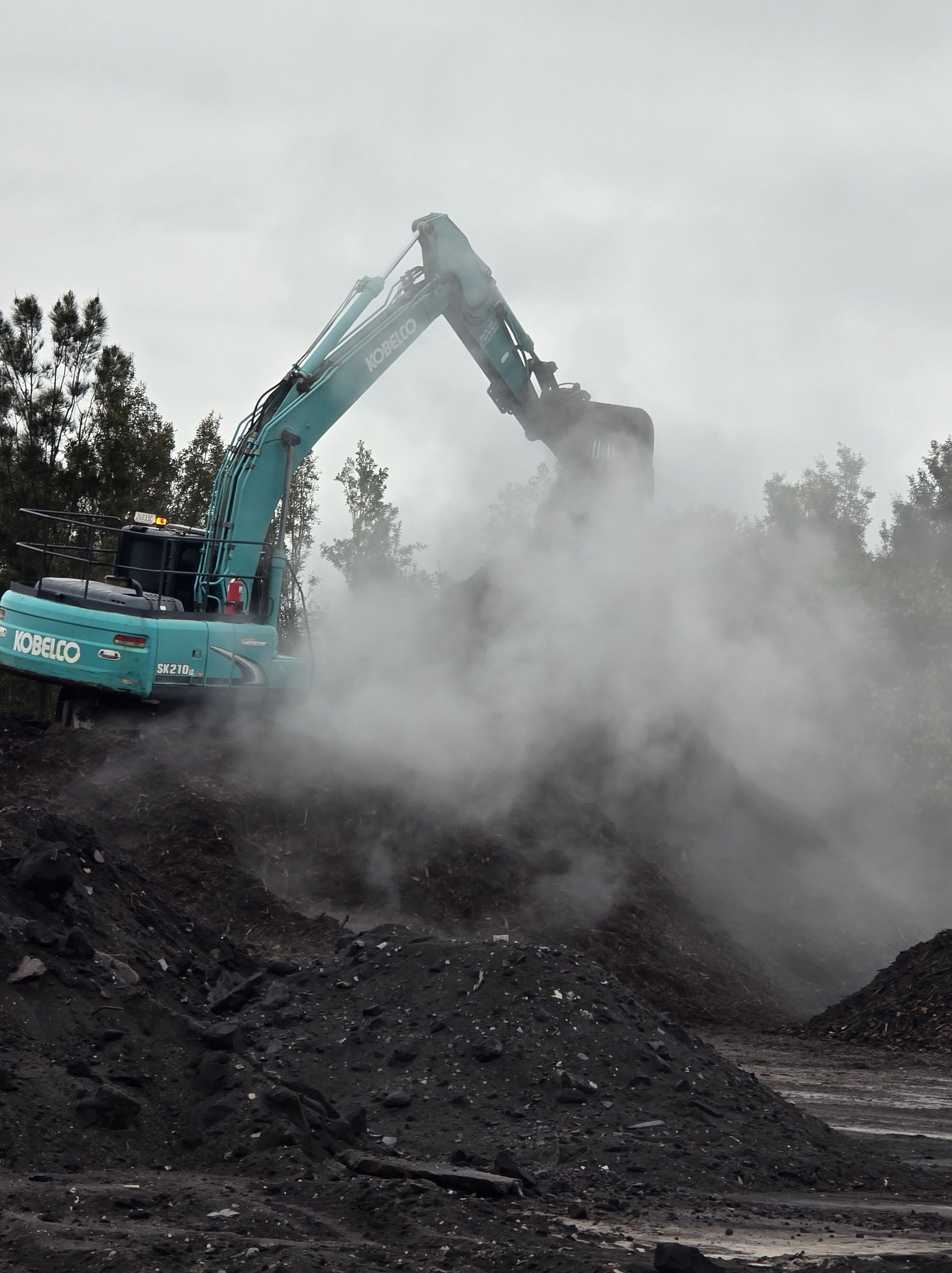 A teal excavator stirs up a cloud of dust on a dark pile of soil — Coastal Sand Soil & Mulch in Craignish, QLD