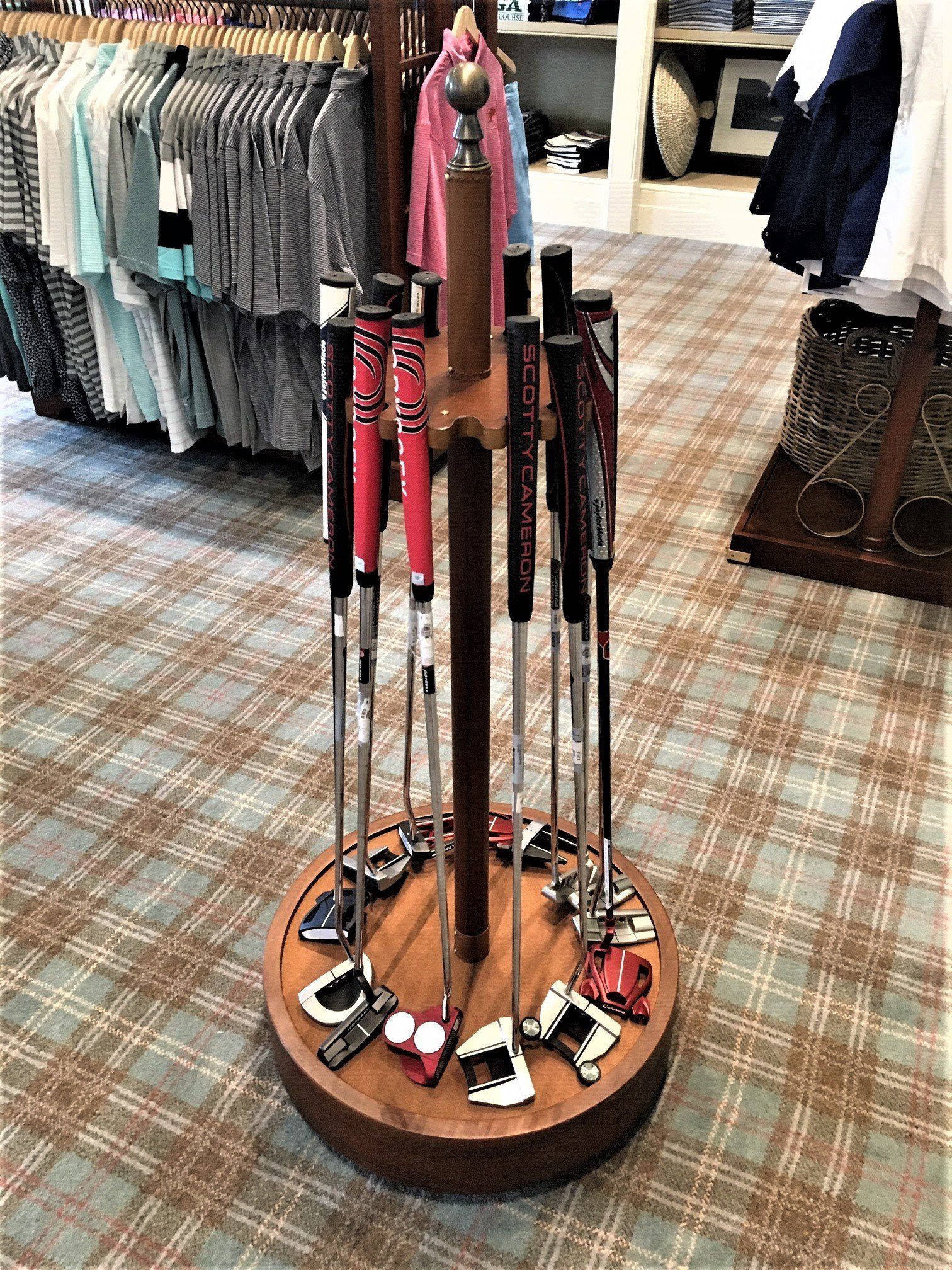 Golf putters displayed in a circular wooden stand on a plaid floor, clothes on hangers in the background.