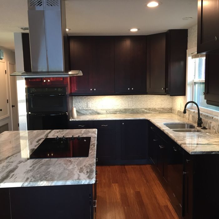 A modern kitchen featuring dark cabinets, light marble countertops, a center island with a cooktop, and wood flooring.
