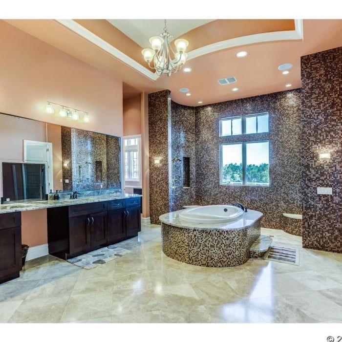 A spacious primary bathroom with a mosaic-tiled oval soaking tub, dark wood cabinets, a large vanity mirror, and chandelier.