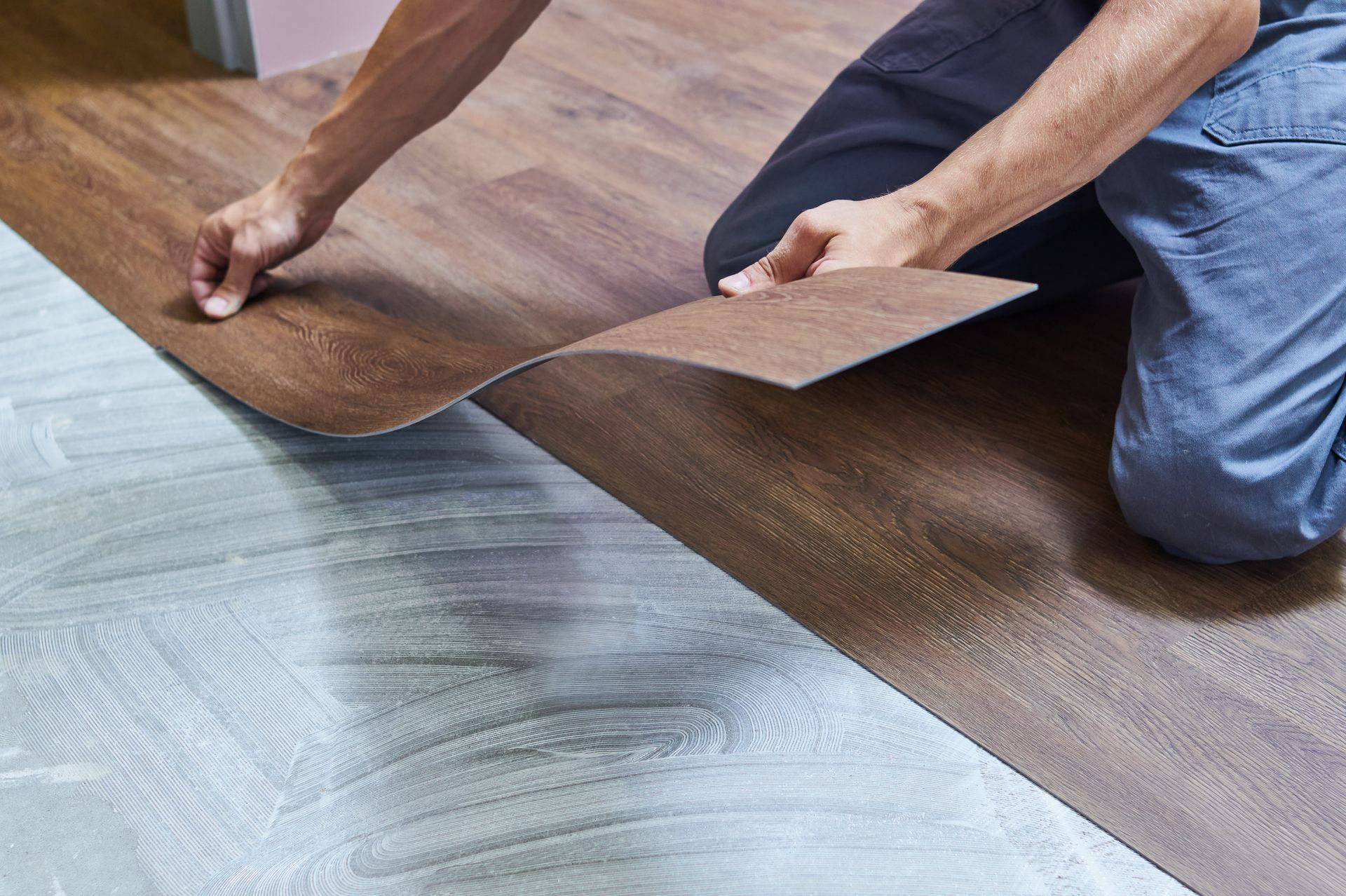 A person installing wood-look vinyl flooring planks onto a white adhesive-covered floor.