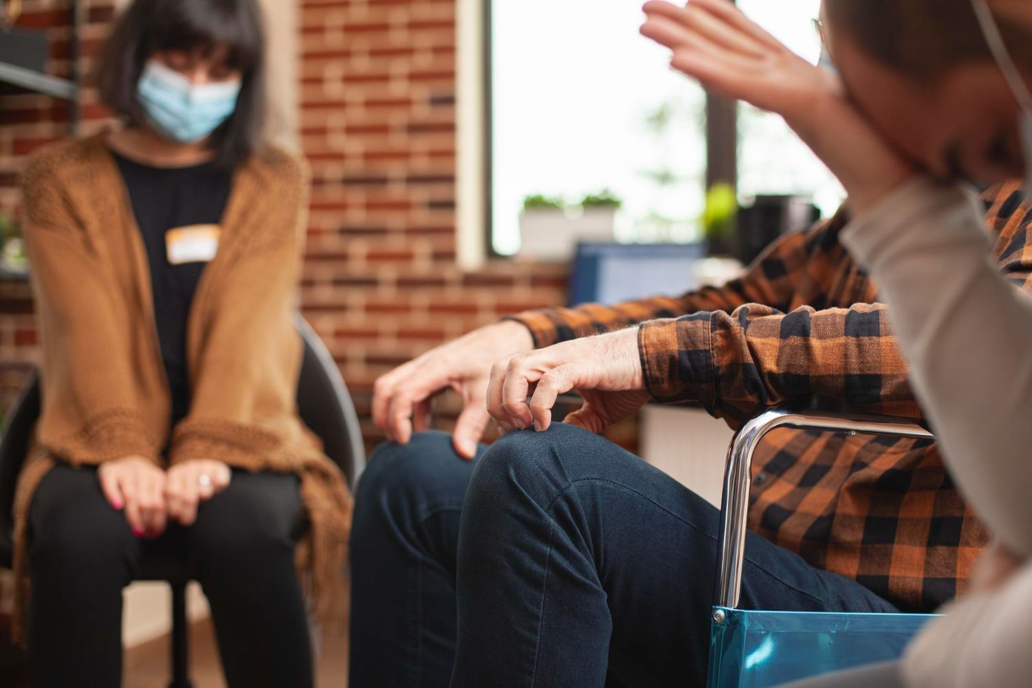 Therapy session with three people. One wears a mask, another gestures with hands, a third hides their face.