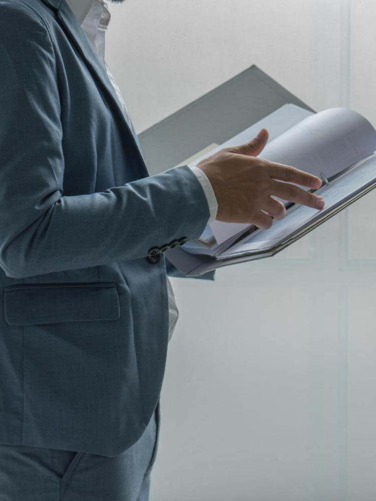 A person in a light blue suit holds a binder and flips through papers against a plain white background.
