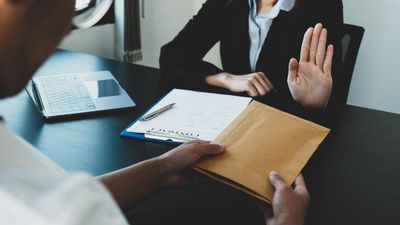 Person offering brown envelope to someone who is refusing with hand up. Office setting.