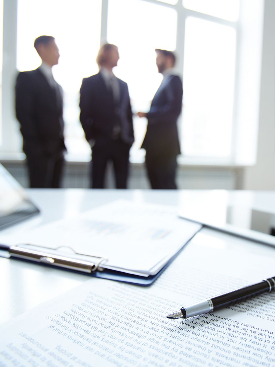 A document and pen on a table, three blurred figures in suits stand in a bright office.
