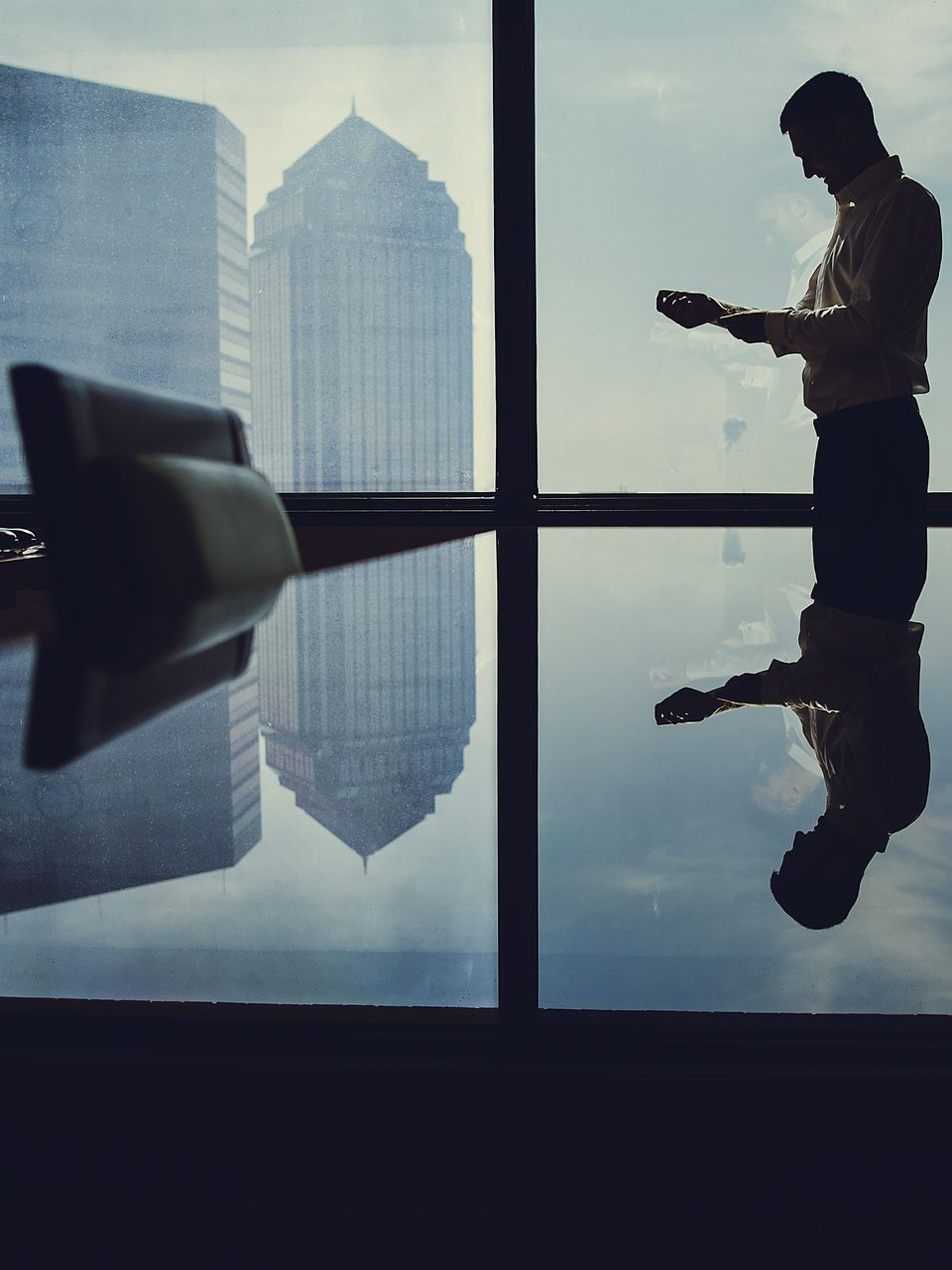 A silhouette of a person standing by a large office window, reflecting on a polished table with city buildings in view.