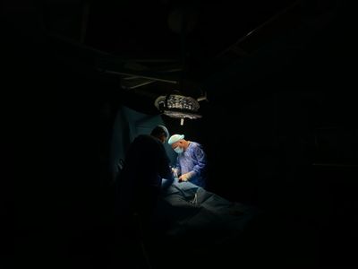 Surgeons operating under a bright light in a dark room. Surgical drapes and equipment visible.