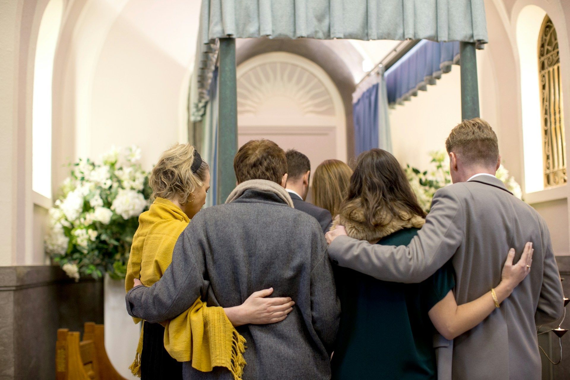 People gathered, embracing, near a covered doorway with a light-blue curtain, likely in a funeral home.