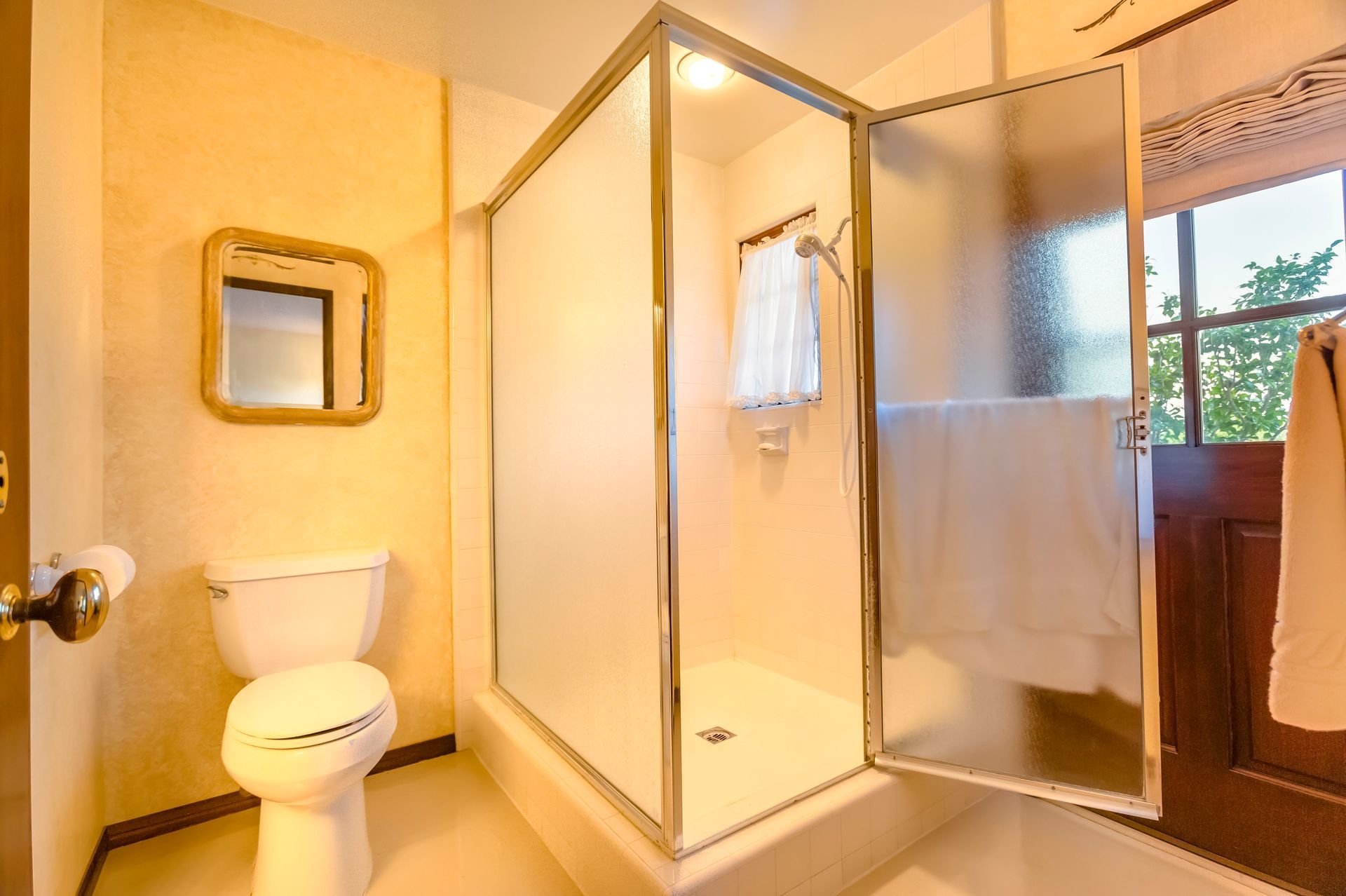 Bathroom with frosted glass shower, toilet, mirror, and doorway. Light walls, towel on shower.