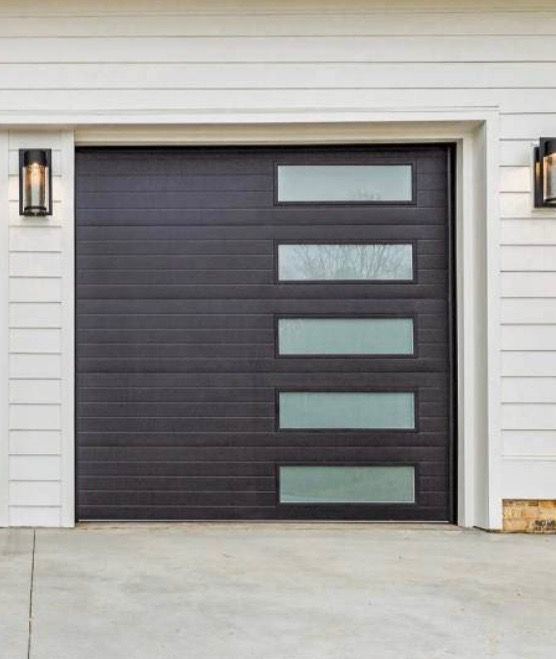 Dark brown garage door with horizontal lines and five rectangular glass panels; flanked by white siding and outdoor lights.