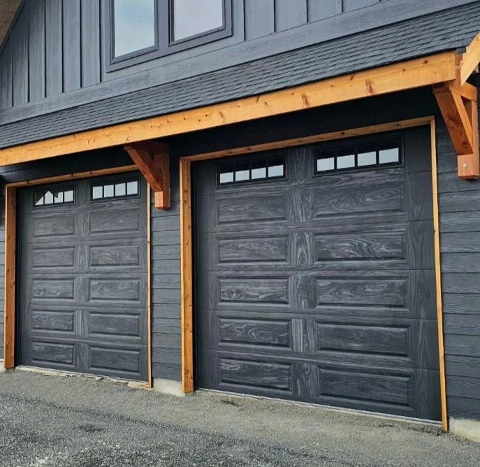 Two dark gray garage doors with wood trim and small glass windows, on a house with dark gray siding.