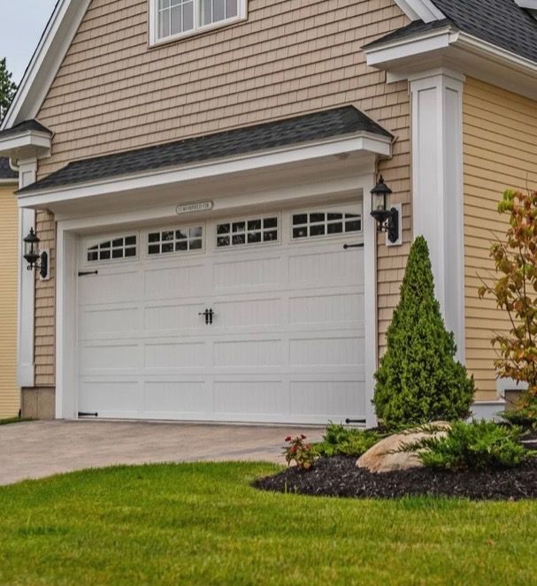 White garage door on a tan house with green lawn, shrubs, and a paved driveway.