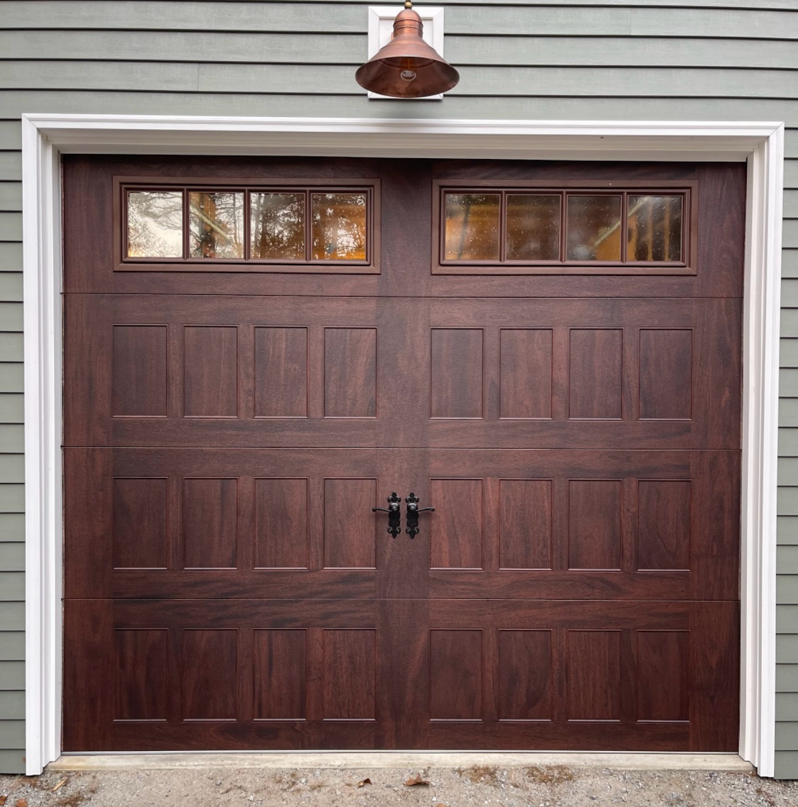 Dark brown garage door with two window panels above, mounted on a building with green siding and a copper light.